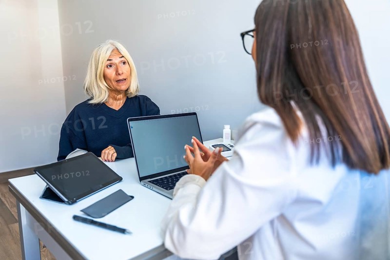Senior woman is talking with a female doctor during a medical consultation in a modern office, discussing her health and treatment options with a laptop and medical equipment on the desk