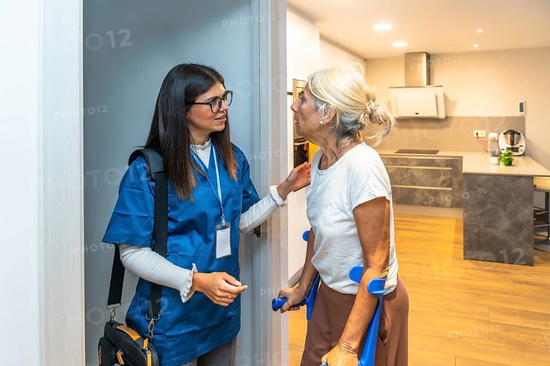 Caregiver wearing scrubs and carrying a medical bag assisting an elderly woman using crutches at the entrance of her brightly lit home, providing personal healthcare support