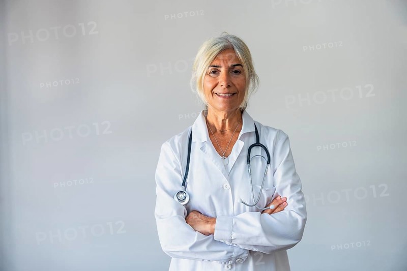 Senior female doctor with gray hair and a lab coat smiling confidently, standing against a neutral background with a stethoscope around her neck, representing healthcare and medical professionalism