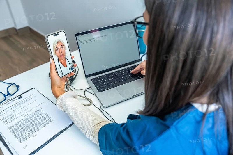 Healthcare professional having a video conference with a doctor on a smartphone while using a laptop, representing modern telehealth services and digital medical communication