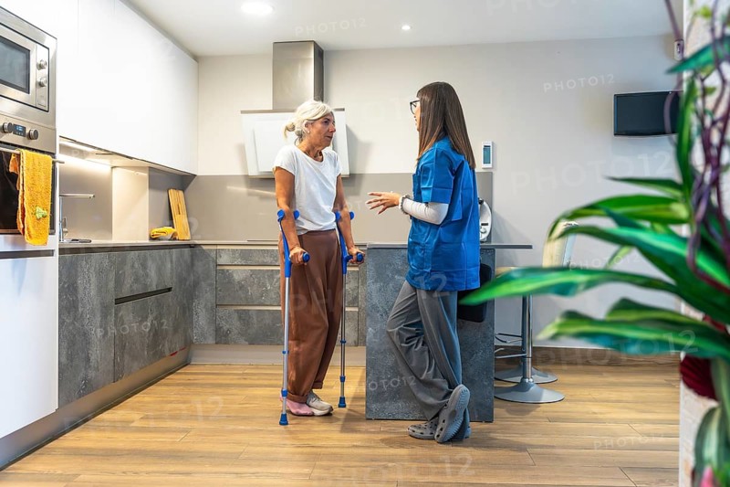 Home caregiver interacting with a senior woman using crutches, providing support and advice for her recovery and rehabilitation process in a modern kitchen setting