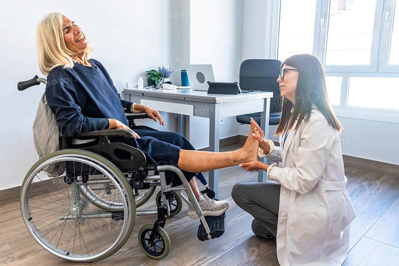 Female doctor examining and rehabilitating a senior woman's leg and ankle, who is seated in a wheelchair and experiencing pain during physical therapy session