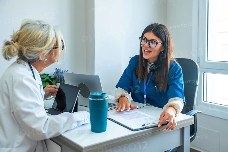 Healthcare professionals, a doctor in a lab coat and a nurse in scrubs, sitting at a desk and discussing medical information during a consultation using digital devices in a clinic office