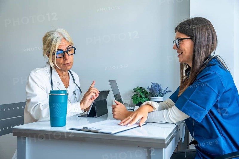 Experienced doctor discussing treatment with a smiling young nurse, both utilizing digital tablets for patient information and modern healthcare collaboration in a clinical office setting