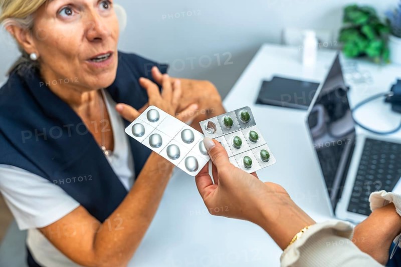 Doctor hand holding two blister packs of pills, one with green capsules and one with silver capsules, offering medicine to a concerned senior woman discussing health issues during a consultation