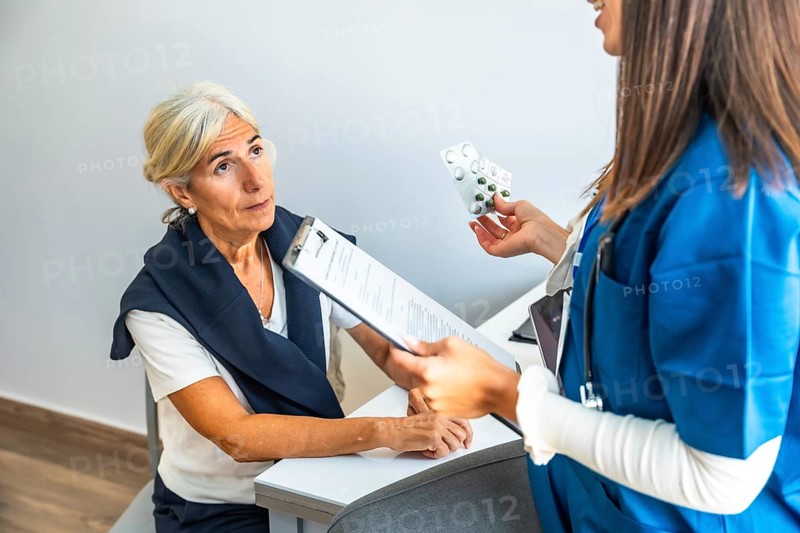 Healthcare professional explaining medication to an attentive elderly patient during annual check up, holding a blister pack of pills and medical forms