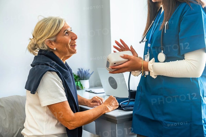 Healthcare professional in blue scrubs holding a blood pressure monitor, explaining its use to an attentive senior woman during a medical consultation