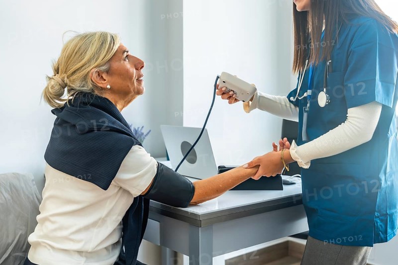 Young medical professional in blue scrubs carefully checking the blood pressure of a smiling senior woman, monitoring her health during a routine checkup in a bright consultation room