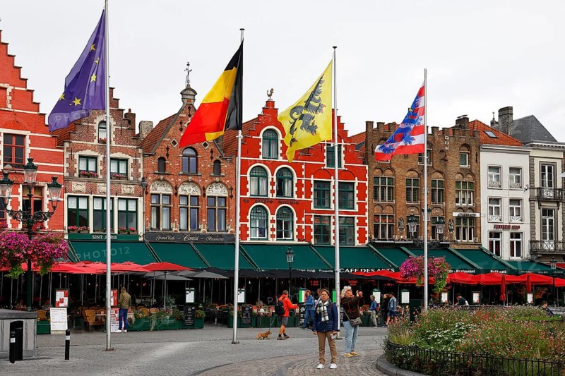 Historic houses on the market square in the old town of Bruges, Grote Markt, former guild houses, now restaurants and cafes, flags of Europe, Belgium, Flanders and Bruges (from left to right), UNESCO World Heritage Site, Flanders, Belgium