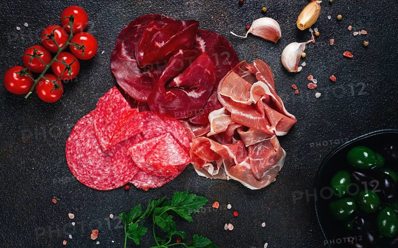 Assorted dried meat, fresh tomatoes and olives spread out on a dark surface, food composition, top view, no people