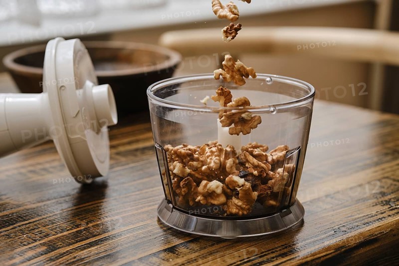 A person is preparing to chop walnuts using a food processor in a cozy kitchen. The walnuts are flying into the container, showcasing an active cooking moment
