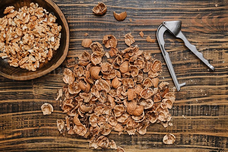 Walnuts are being shelled on a rustic wooden surface. A bowl of walnut pieces sits nearby, while the shells are scattered around, showcasing the process of nut preparation in a cozy kitchen setting