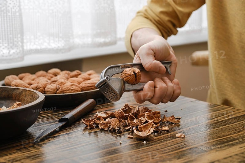 A person is breaking open a walnut with a metal nutcracker, sitting at a wooden table. Shelled nuts are scattered around, creating a warm and inviting atmosphere in the kitchen during daylight hours