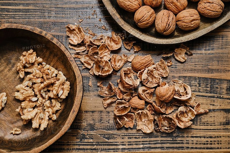 Whole walnuts and broken shells scattered across a wooden table. Some walnut pieces are neatly placed in a separate wooden bowl, showcasing their natural beauty