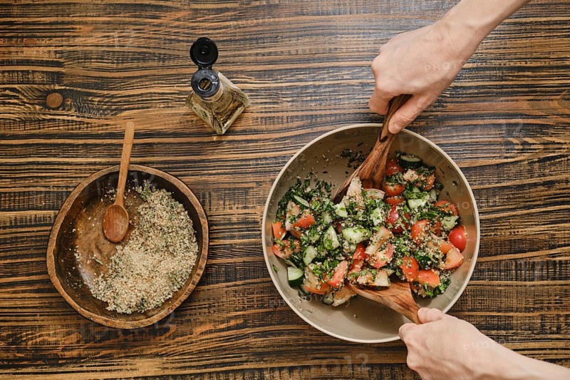 Two hands mix fresh vegetables in a bowl while preparing a salad. Nearby, a smaller bowl holds seasoning and garnishing ingredients, creating a vibrant and tasty dish