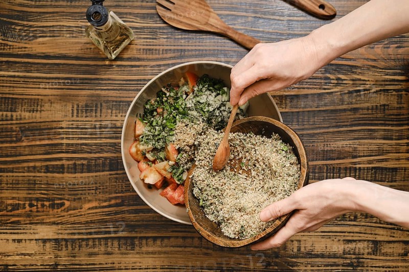 Adding ground walnuts to a bowl with chopped vegetables and herbs. A wooden spoon is used to combine the fresh ingredients. Olive oil and utensils can be seen in the background
