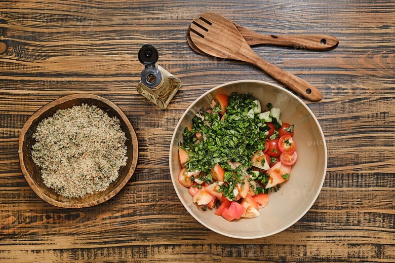 Chopped tomatoes and fresh herbs in a bowl ready for mixing. A bowl with crushed walnuts is nearby, alongside wooden utensils on a rustic wooden table
