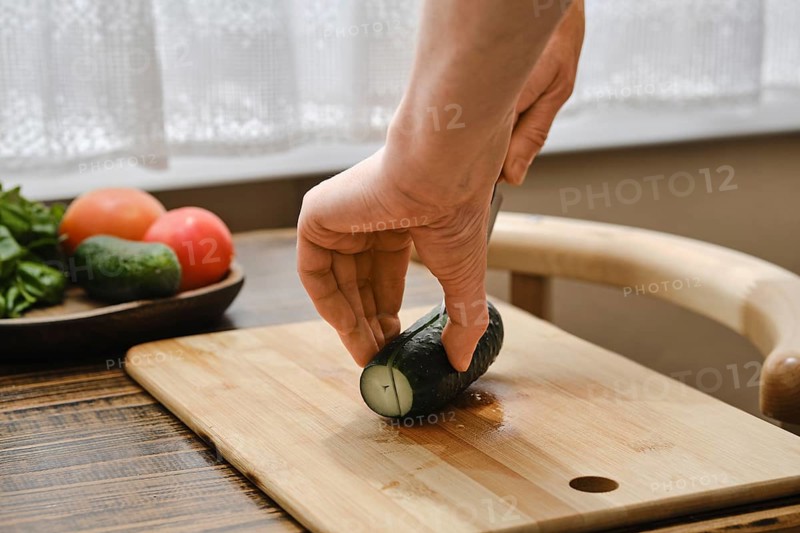A hand holds a cucumber while cutting it on a wooden board. Fresh tomatoes and greens are arranged on a plate beside it, suggesting a meal preparation. Sunlight shines through the window