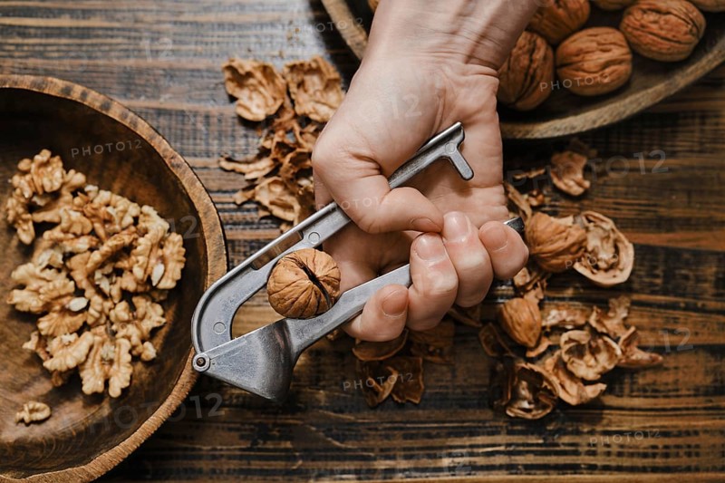 A person is holding a metal nutcracker, squeezing it to crack open a walnut. Chopped walnuts and empty shells surround the tool on a rustic wooden surface, creating a natural, cozy setting