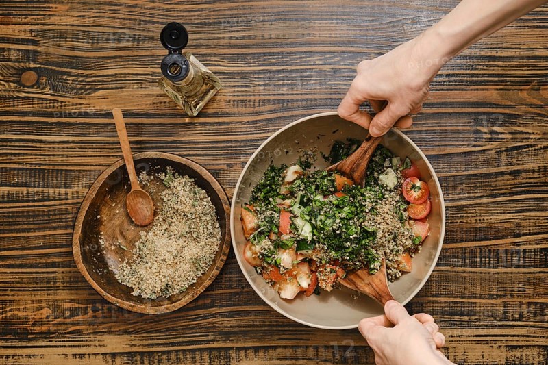 Hands gently mix fresh tomatoes, herbs, and chopped walnuts in a large bowl. Wine vinegar oil is nearby for drizzling over the salad