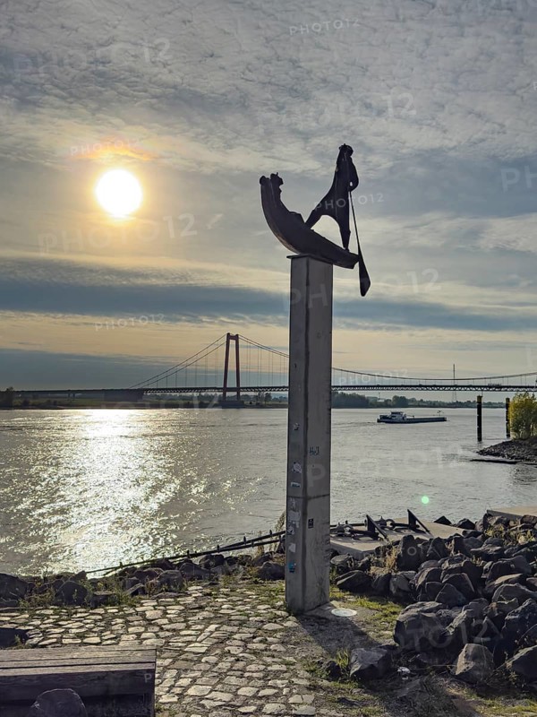 Photo with melancholy atmosphere of sculpture Fährmann Artwork on pillar stele by sculptor Dieter von Levetzow from 1983 on the Rhine promenade in Emmerich, reminiscence of time in front of the Rhine bridge, on the river right under Rhine bridge Freight ship freighter for inland navigation, Emmerich, Lower Rhine, North Rhine-Westphalia, Germany