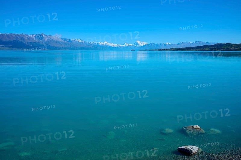 Clear, calm lake in front of a distant mountain range under blue sky, Lake Pukaki Viewpoint, Pukaki, Canterbury, South Island, New Zealand