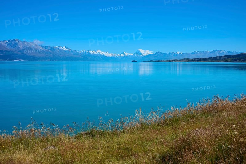 View over grassy banks to the mountain range behind the lake, Lake Pukaki Viewpoint, Pukaki, Canterbury, South Island, New Zealand