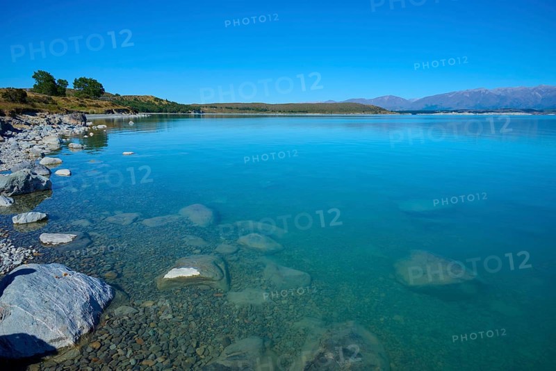 Shore with stones in clear water surrounded by vast countryside and mountains, Lake Pukaki Viewpoint, Pukaki, Canterbury, South Island, New Zealand