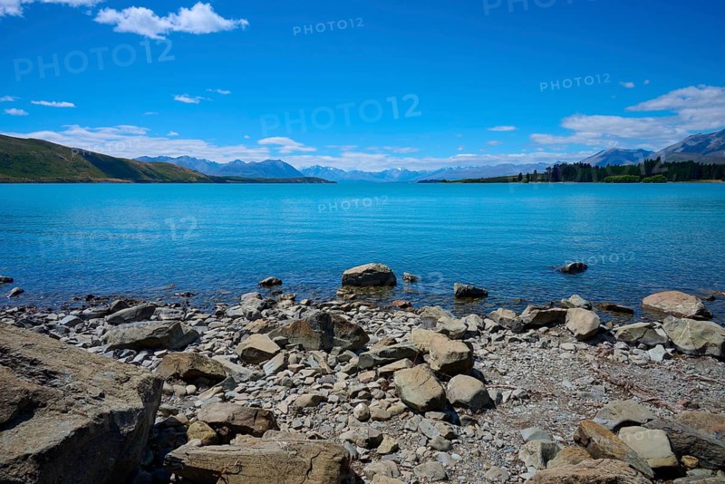 Clear water gently washes against rocky shoreline surrounded by high mountains under bright blue skies, Lake Tekapo, Tekapo, Canterbury, South Island, New Zealand