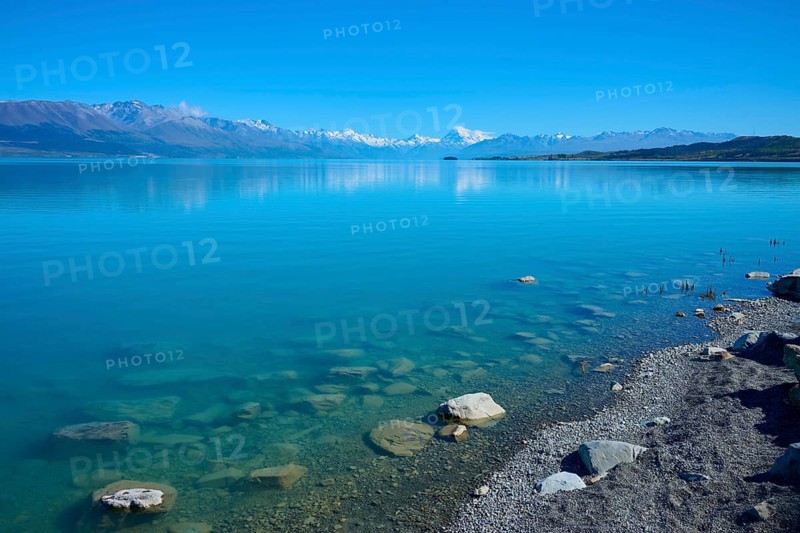 Clear blue water with rocks on the shore and snowy mountains in the background, Lake Pukaki Viewpoint, Pukaki, Canterbury, South Island, New Zealand