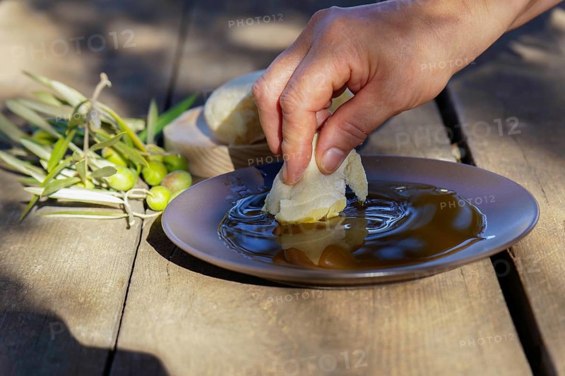 Hand dipping a piece of fresh bread into a shallow plate of extra virgin olive oil, with olives and olive branches on a rustic wooden table