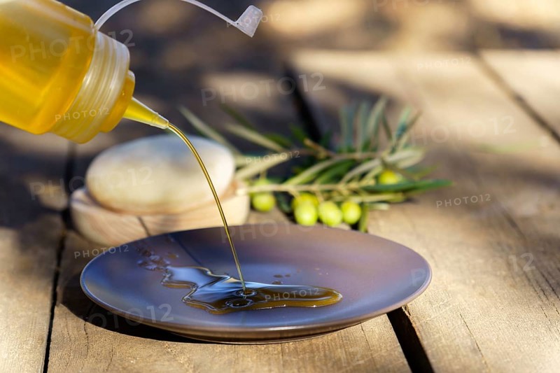 Pouring fresh extra virgin olive oil from a squirt bottle onto a plate, with olive branches in the background