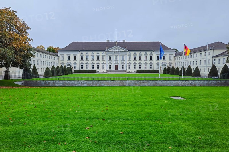 Neoclassical castle in front of a well-kept lawn, lined with topiaries, under grey skies with waving flags and a quiet autumn atmosphere, at Bellevue Palace in 10557 Berlin, Germany