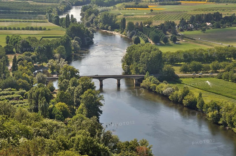 An aerial view of a serene river flowing under a bridge, surrounded by lush green fields and trees. Dordogne seen from Domme in Périgord, France