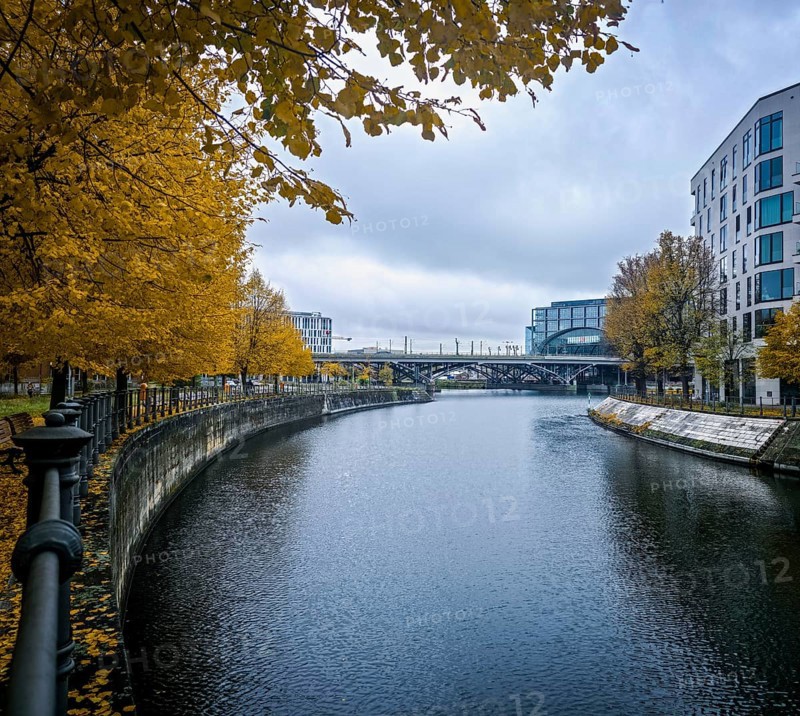 View along a quiet canal, lined with glowing yellow autumn trees and modern architecture, under a cloudy sky, at Berlin Central Station, 10557 Berlin, Germany