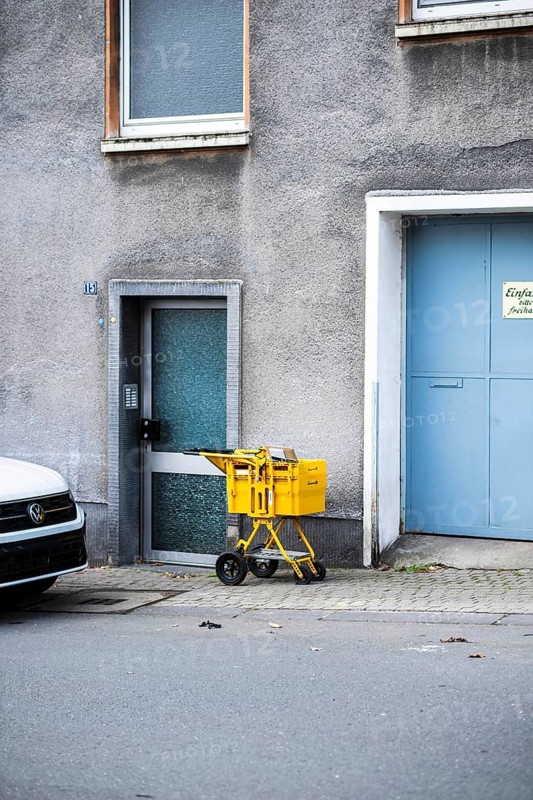 A yellow Deutsche Post delivery van stands in front of a grey façade between a front door and a large, light blue garage door, Wuppertal, Germany