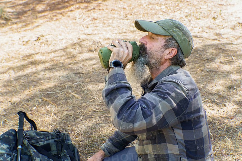 Mature bearded hiker drinking from a canteen during a trekking break in sunlit forest, resting and rehydrating on trail (outdoors)