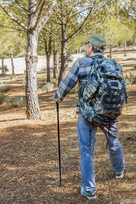 Man walking an outdoor path, carrying a camouflage backpack and trekking pole, enjoying nature and adventure