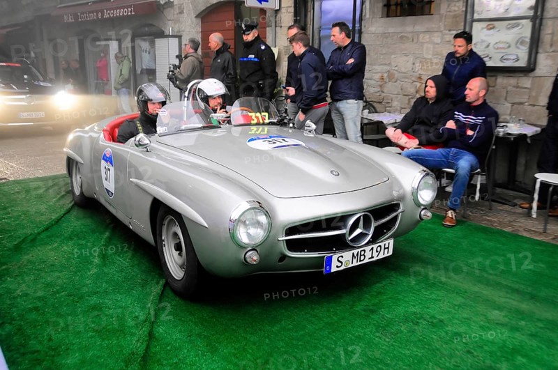 Silver 190 SL Roadster with helmeted occupants slowly across the green carpet, watched by spectators in front of stone facades, Mille Miglia 2016, time control, checkpoint, SAN MARINO, classic car racing. San Marino, Italia