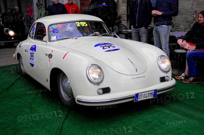 White Porsche 356 with glowing headlights drives across green carpet during a vintage car rally, watching passers-by, Mille Miglia 2016, time control, checkpoint, SAN MARINO, classic car racing. San Marino, Italia