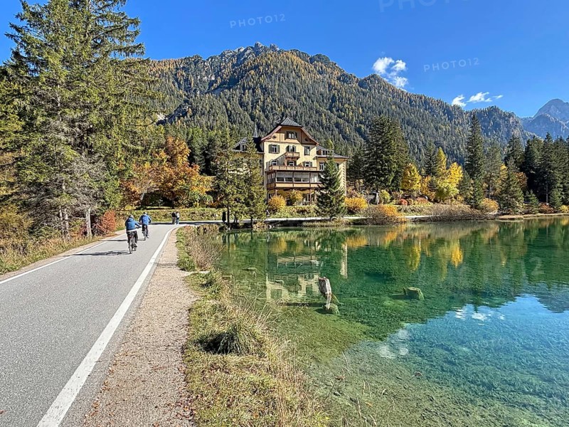 Idyllic autumn scene with a clear lake surrounded by trees and mountains, cyclists on a narrow road, cycle tourism, Lake Toblach, Lake Dobbiaco, South Tyrol, Italy