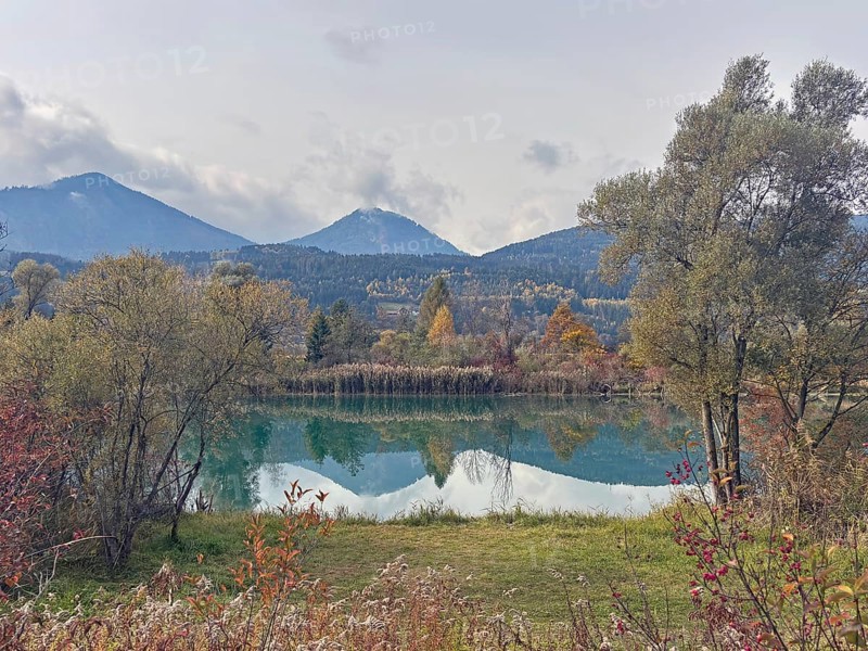 A lake in autumn surroundings, surrounded by mountains and trees under an overcast sky, Drau, Villach, Drauradweg, Carinthia, Austria
