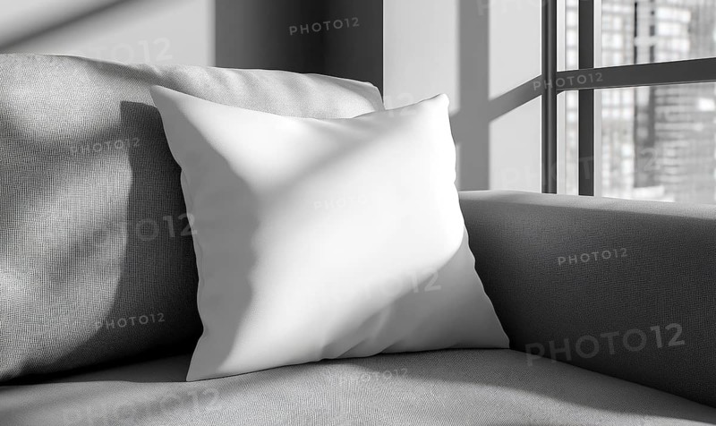 A white pillow sits on a gray couch in front of a window. The pillow is placed on the couch in such a way that it is almost as if it is floating. AI generated