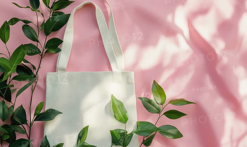 A white tote bag is placed on a pink background with green leaves. The bag is empty and the leaves are in the foreground. Concept of simplicity and natural beauty. AI generated