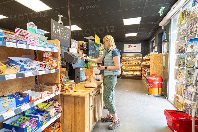 A woman pays at a supermarket checkout, surrounded by shelves of snacks and products, 24 hours Tante M Store
