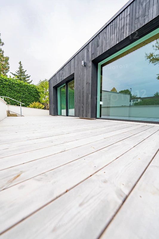 Deep view along the wide wooden terrace of the modern, black-clad building with floor-to-ceiling windows, house renovation, air-conditioning renovation, Metzingen, Germany