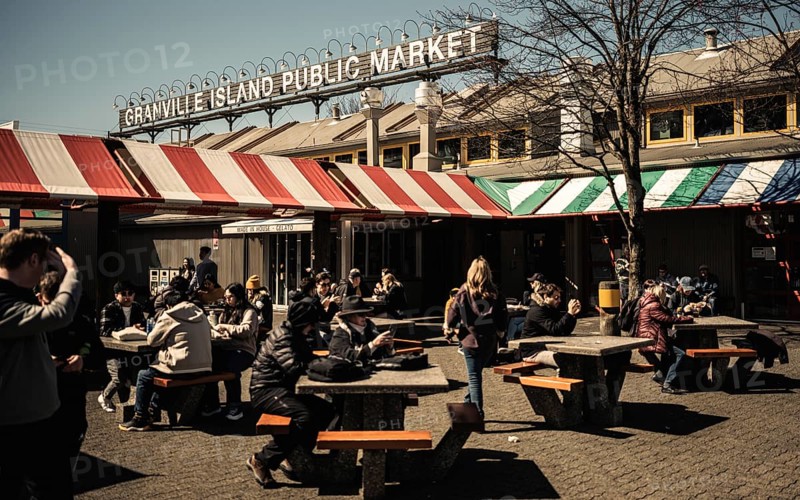Crowds enjoy a lively day at an outdoor market under warm sunlight on Granville Island, in Vancouver, British Columbia, Canada