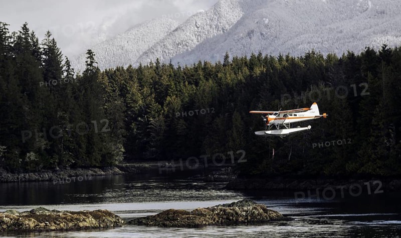 A seaplane flies above rocky shores and dense forest with snow-capped mountains in the background in Tofino, British Columbia, Canada