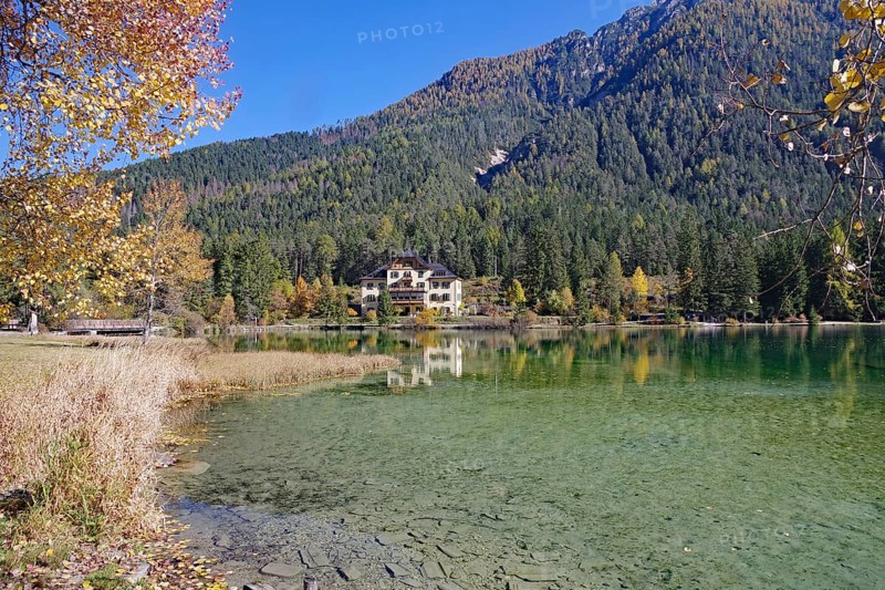 Wide shore with shallow, crystal-clear water, opposite is a villa in the sunshine in autumn forest, Lake Toblach, Lake Dobbiaco, South Tyrol, Italy