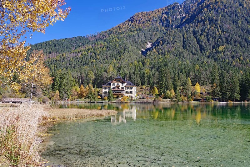 Clear shore water of an alpine lake, opposite is a villa reflected against colorful autumn forests under a deep blue sky, Lake Toblach, Lake Dobbiaco, South Tyrol, Italy
